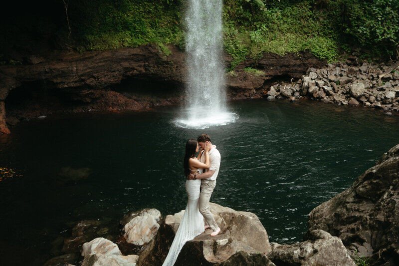 Elopement Photo Ideas | Eloping couple standing on a big rock with a huge waterfall behind them perfectly lined up with their bodies 