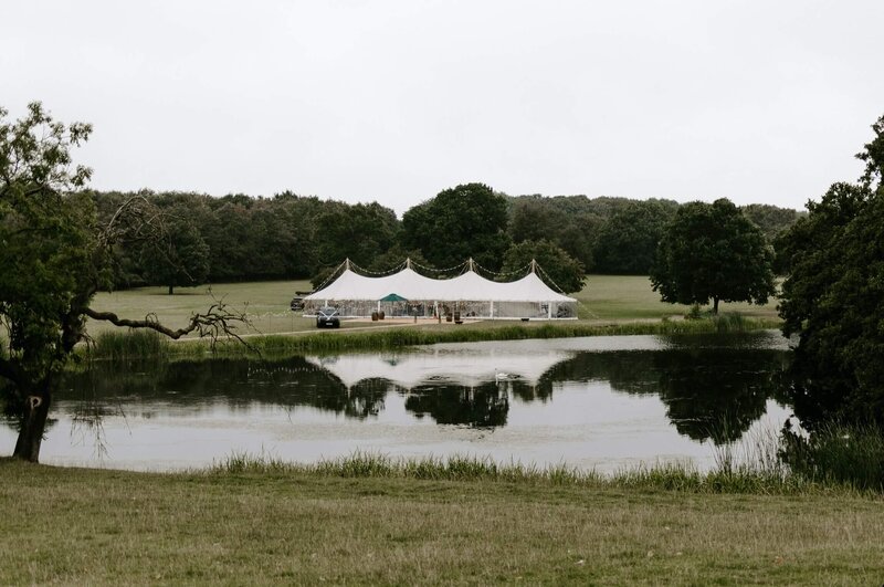 A large pole marquee in a field behind a lake with stunning country views