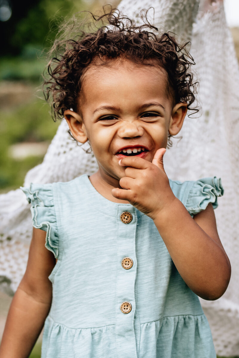 A close-up of a cheerful toddler with curly hair, wearing a light blue dress, smiling and playfully putting a finger in their mouth.