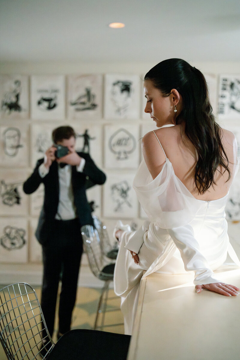 Bride sitting on counter in modern art-filled room while groom photographs her on film