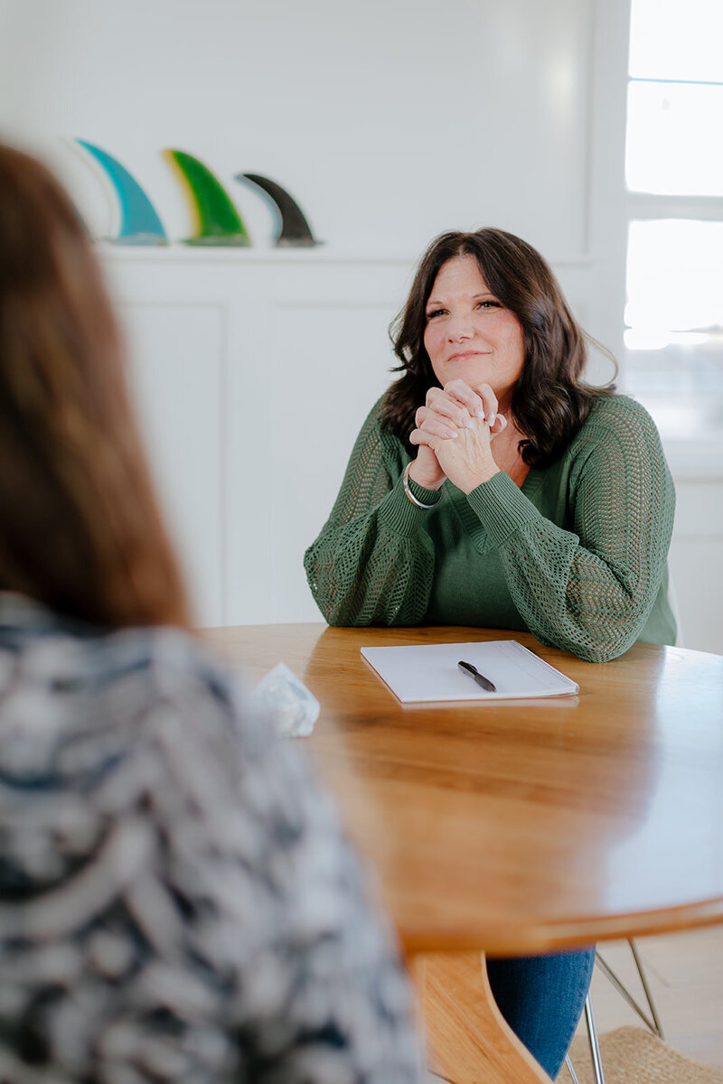 Susie Schumacher smiling warmly during a coaching conversation at a round wooden table, wearing a green sweater with a notepad and pen in front of her - Susie Schumacher Life Coach