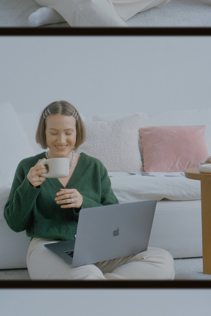Margarita sits on the floor, leaning against a white couch behind her, her lapton on her lap, and.a smooth, cream coloured teacup in her hand. She smiles jovially while reaching for another sip of her matcha, which mathces her grassy green sweater.