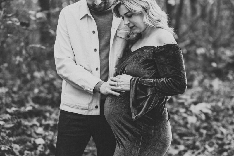 A maternity photo depicting a pregnant woman wearing a dark velvet dress, standing outdoors. She is cradling her belly with one hand while her partner, dressed in a light jacket, places his hand on her belly, signifying affection and support.