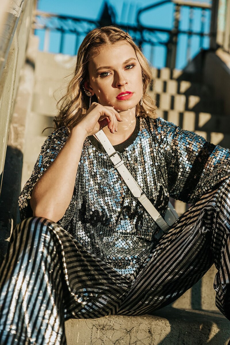 Confident model in a silver sequin top and striped pants, seated on concrete steps in golden sunlight during a gritty-glam workshop session with Tara Rochelle.