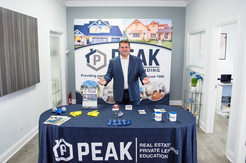 A man smiling with his arms open standing in front of the Peak Private Lending exhibitor setup.