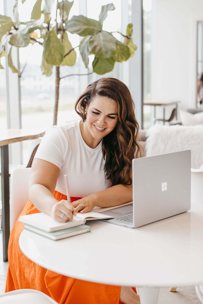 Woman with dark hair, white shirt, orange skirt on laptop and journaling