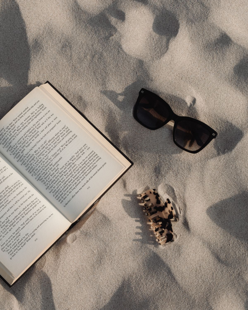 n open book, sunglasses, and seashells arranged on warm sand under golden sunlight — capturing a tranquil moment of beach reading.