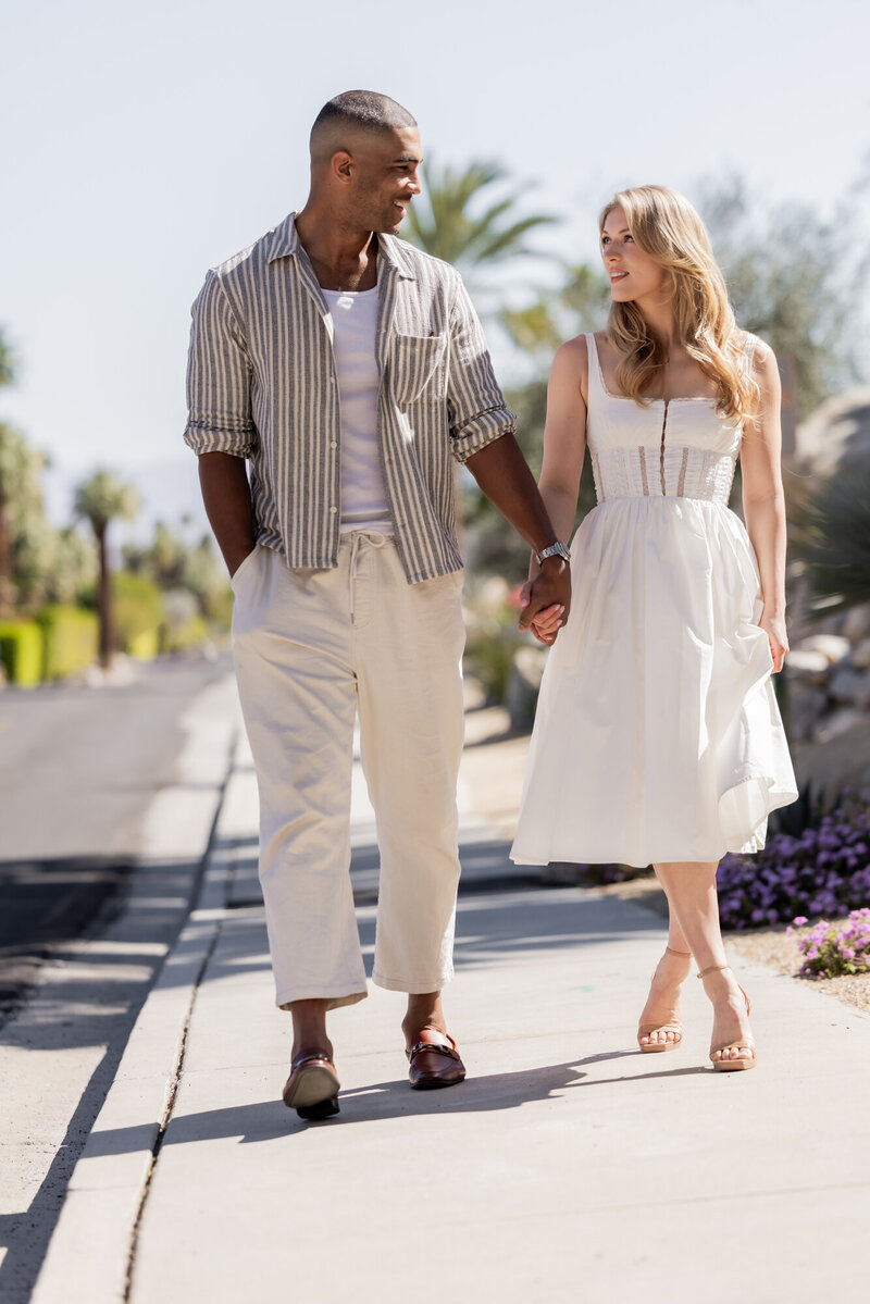 Couple walking through a garden pathway in soft light, holding hands and smiling in relaxed, romantic fashion.