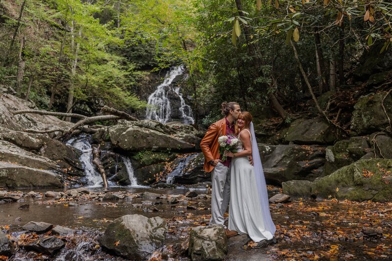A bride and groom stand in front of a large, tumbling waterfall. The groom is kissing the brides head while the bride is smiling happily at the camera. The bride is in a flowy wedding dress with a long veil and the groom is wearing tan pants with an orange leather jacket. They are surrounded by rocks and lush green trees.