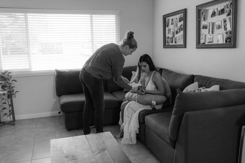 A woman is sitting on a couch in a living room while trying to nurse her newborn, while her partner helps