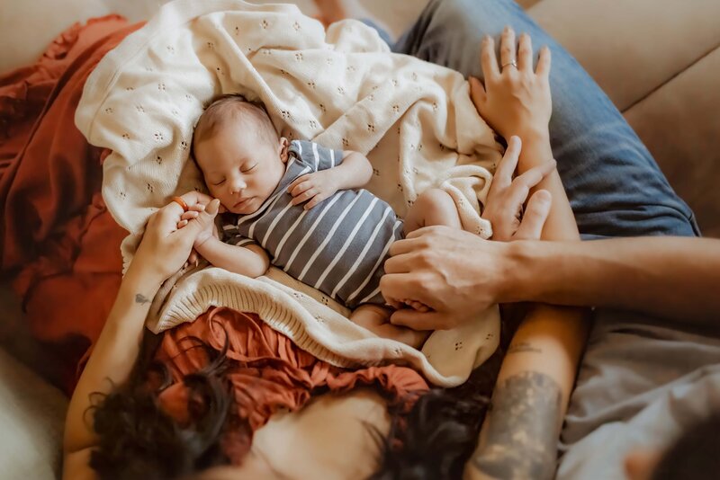 A newborn baby, dressed in a striped onesie, sleeps on a cozy blanket while being cradled by two adults, one wearing a red shirt and the other in blue jeans, with tattoos visible on their arms.