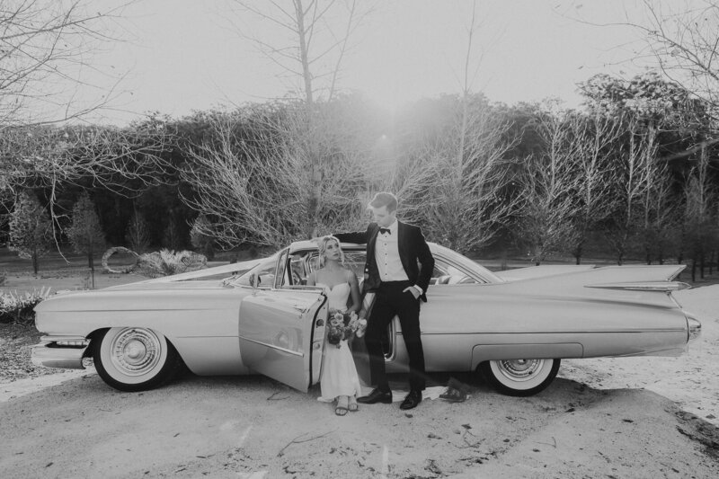 A couple looking cool and sophisticated at their Elopement wedding. They are sitting in a classic cadillac and have bright bold pink flowers and veil