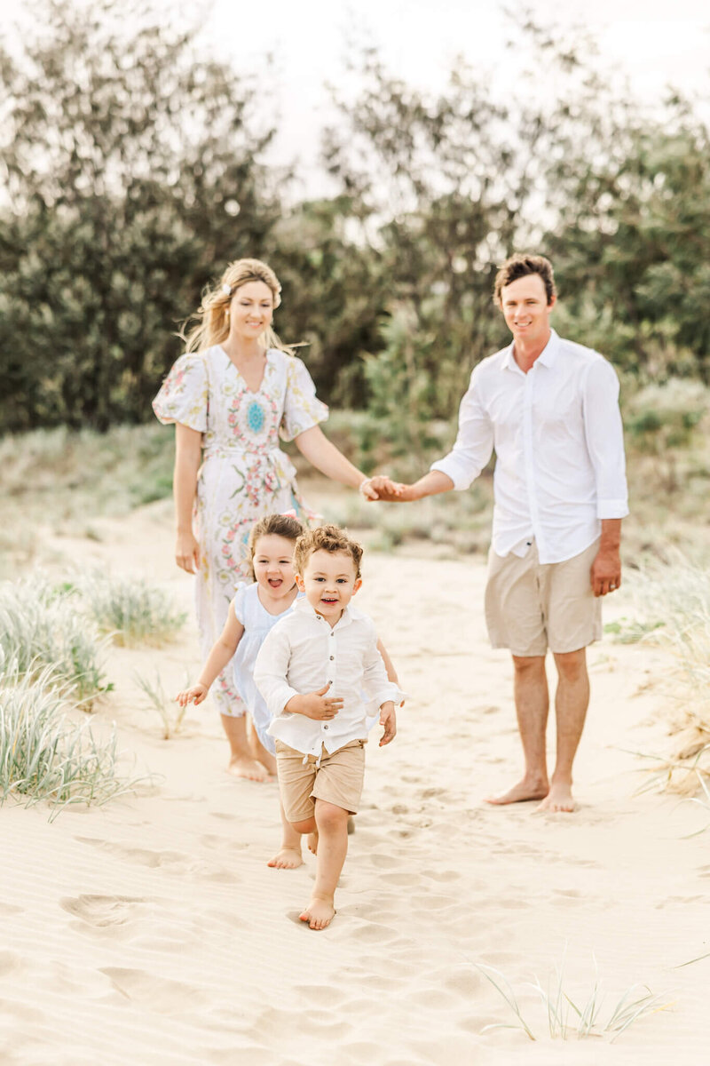 Family portraits of mum and dad holding hands while kids run ahead at the beach.