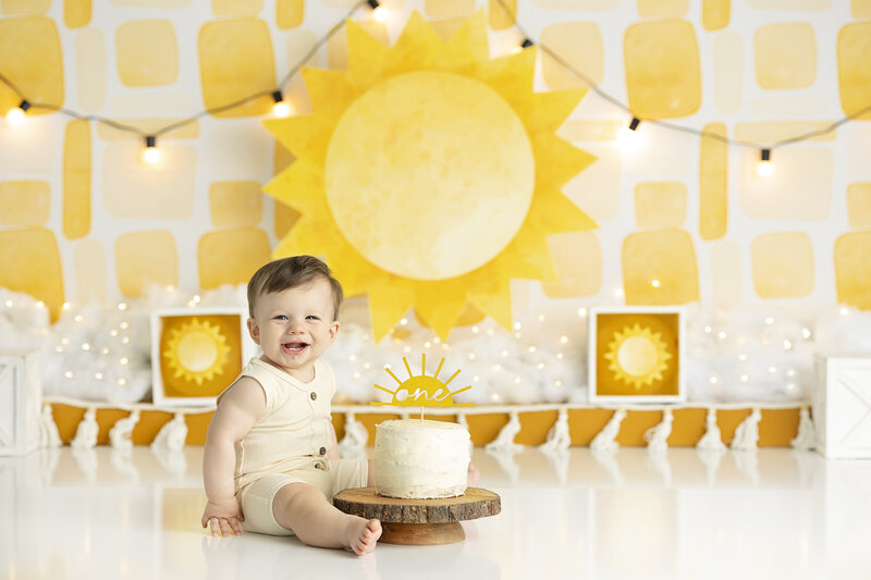 1 year old having fun in a cake smash session sitting on yellow backdrop