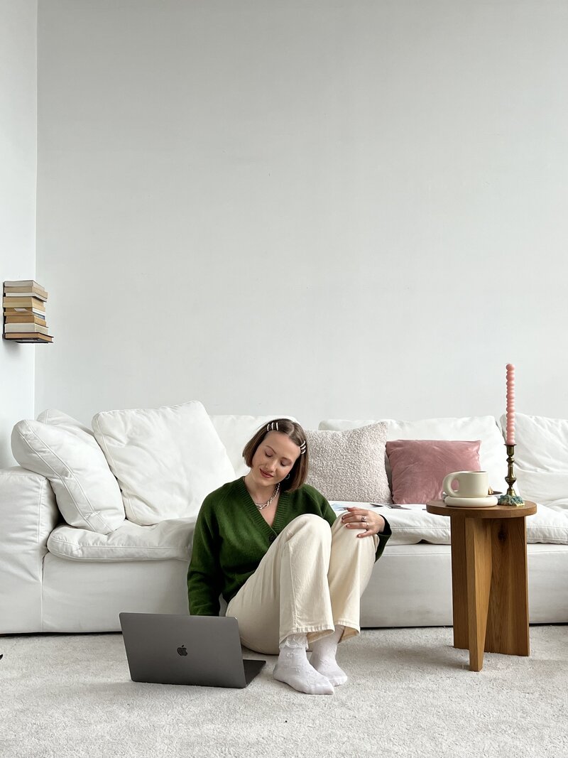Margarita sitting on the floor with her laptop on one side, scrolling and smiling at the screen. A wooden table beside her. A big white couch behind her.