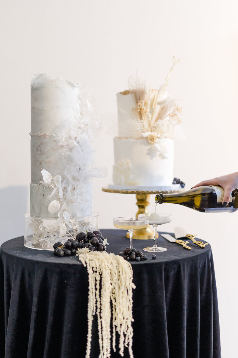 2 and 3 tiered modern wedding cakes on a black velvet linen round high-top table with gold cake cutlery and two ribbed champagne coupes with a hand pouring champagne into the coupes