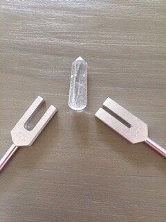  Two tuning forks are positioned on either side of a clear quartz crystal wand on a wooden surface, representing sound healing or energy work.