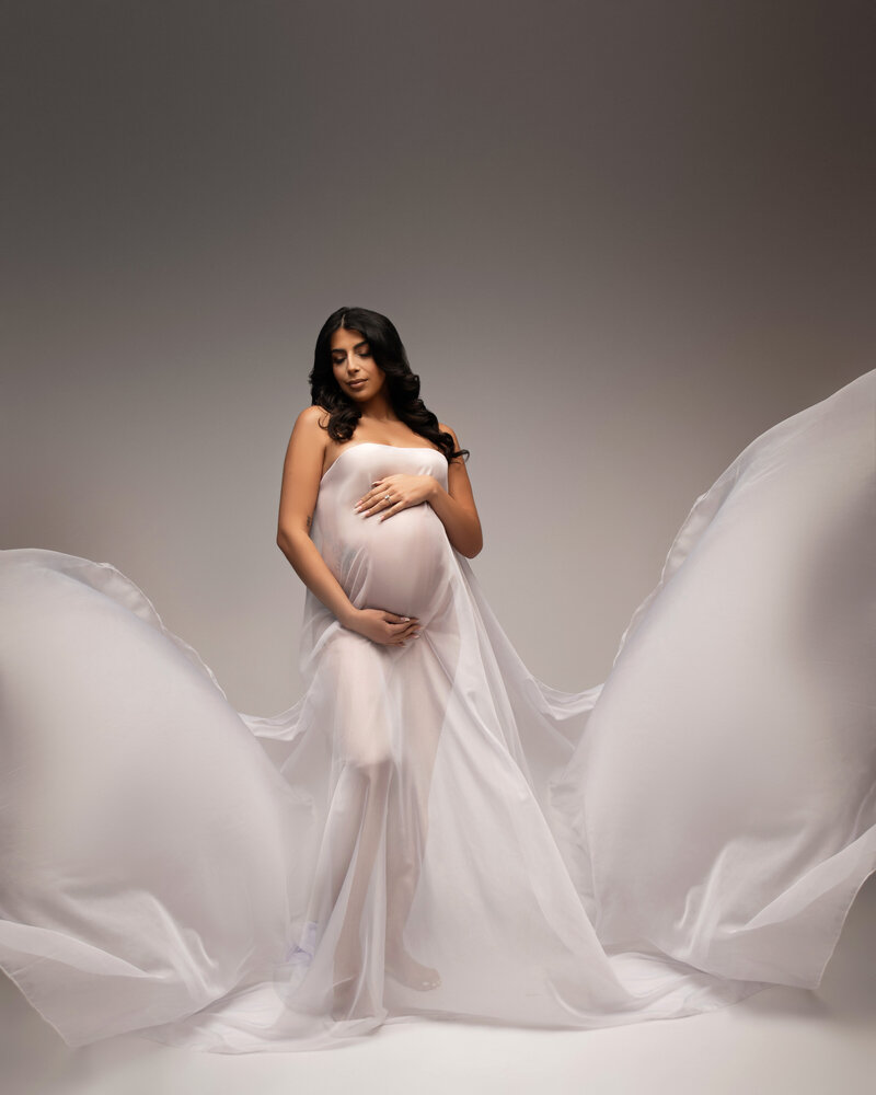 Beautiful pregnant woman with black hair draped in white flowing fabric on a grey backdrop with eyes closed