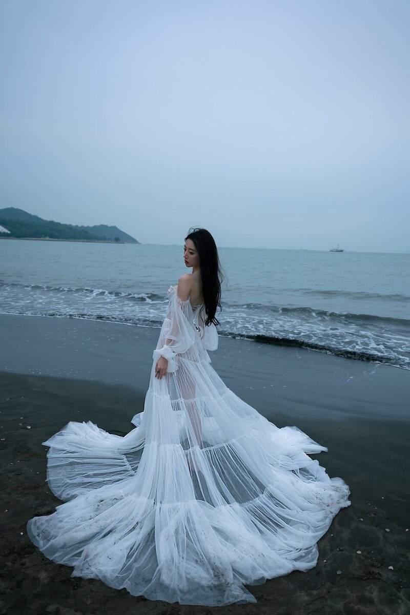 A woman wearing a delicate sheer couture dress shot on the beach in dark moody tones.