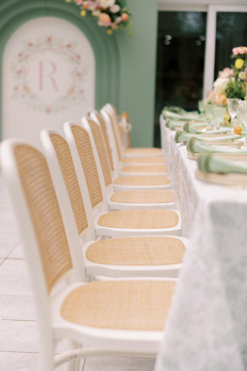Close-up row of white cane-back chairs along a long table set with pastel florals and green napkins.