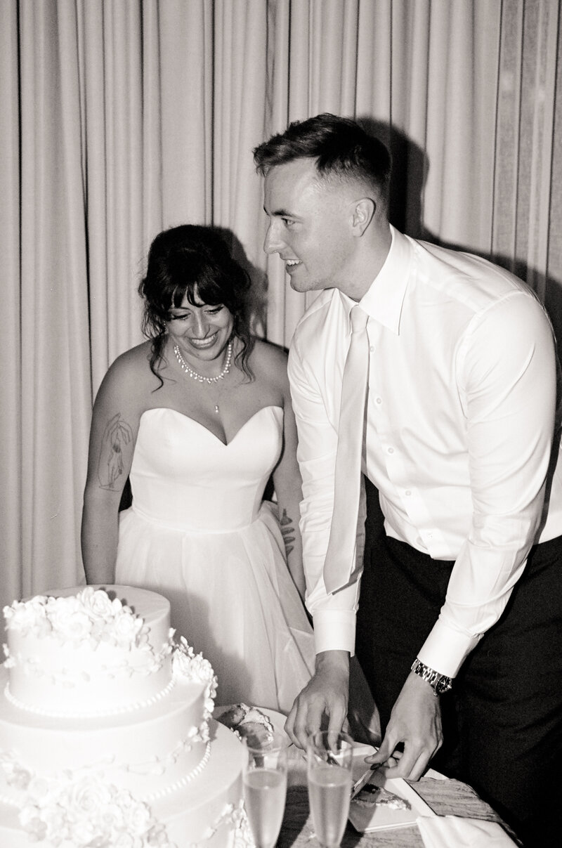 Black and white film photo of a bride and groom smiling while cutting their cake during a summer wedding at the Kimpton Marlowe Hotel in Cambridge, MA — a sweet moment made by a film wedding photographer.