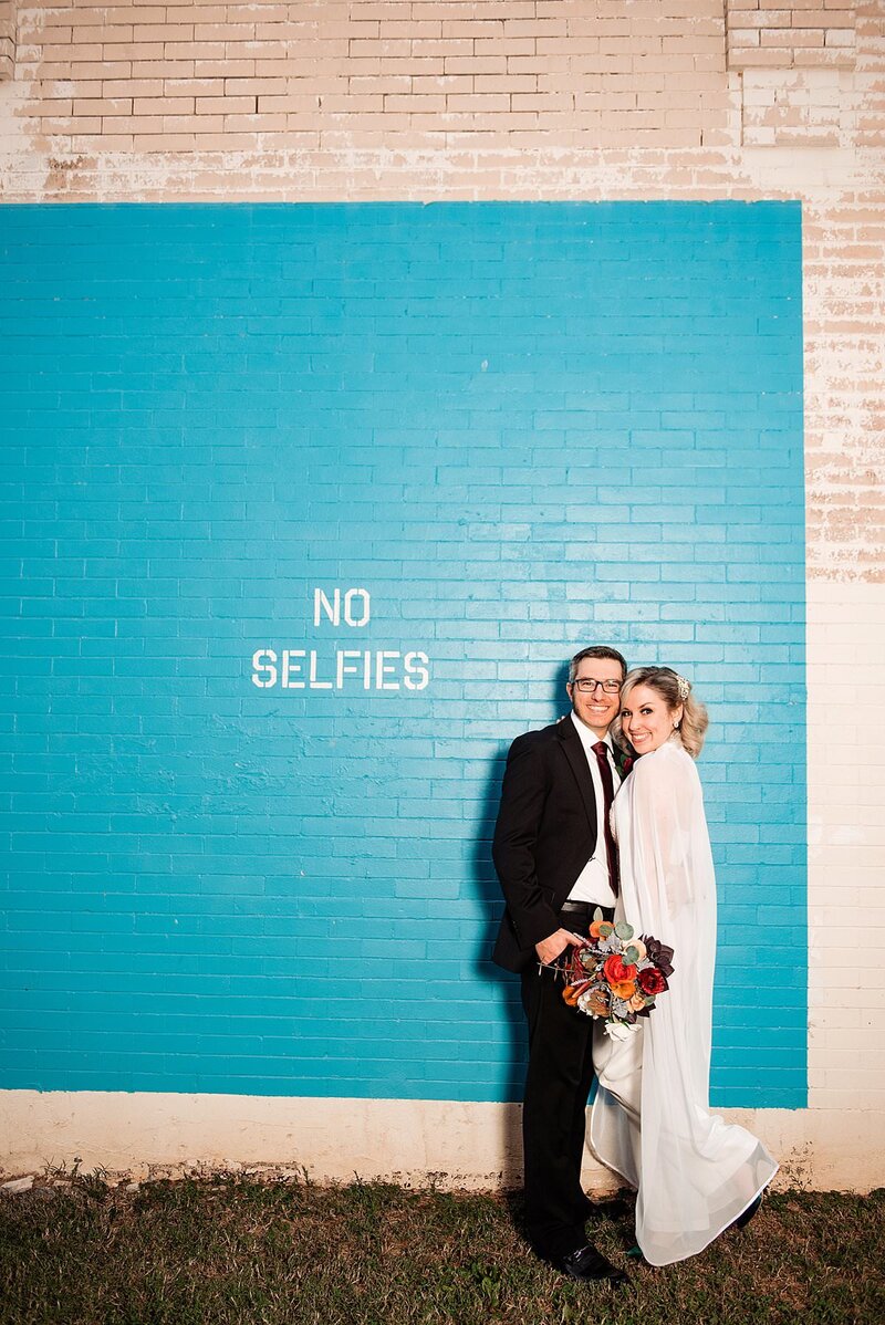 Couple standing together against a wall with a No Selfies mural, they are in their wedding attire and smiling at the camera