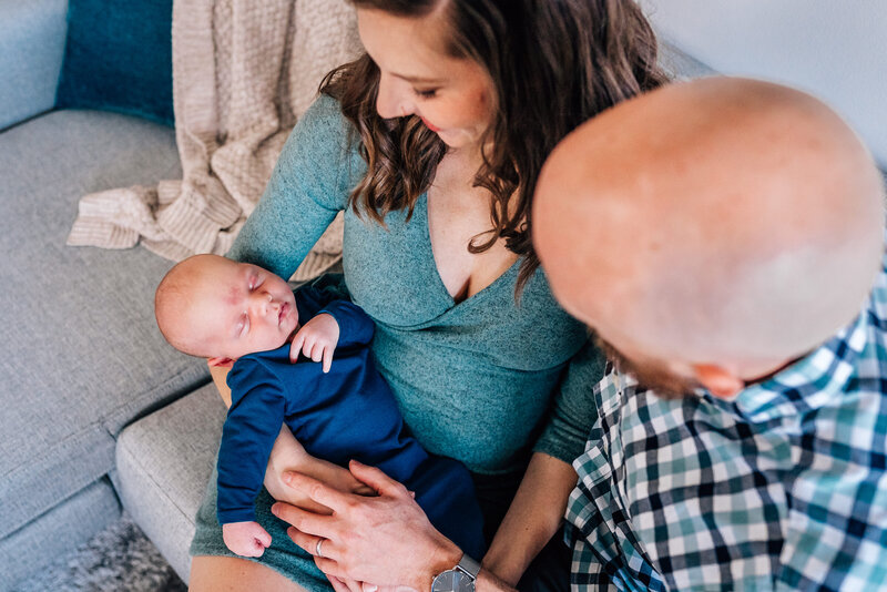 A mother and father sit together on a couch, admiring their sleeping newborn baby who is dressed in a cozy blue outfit.