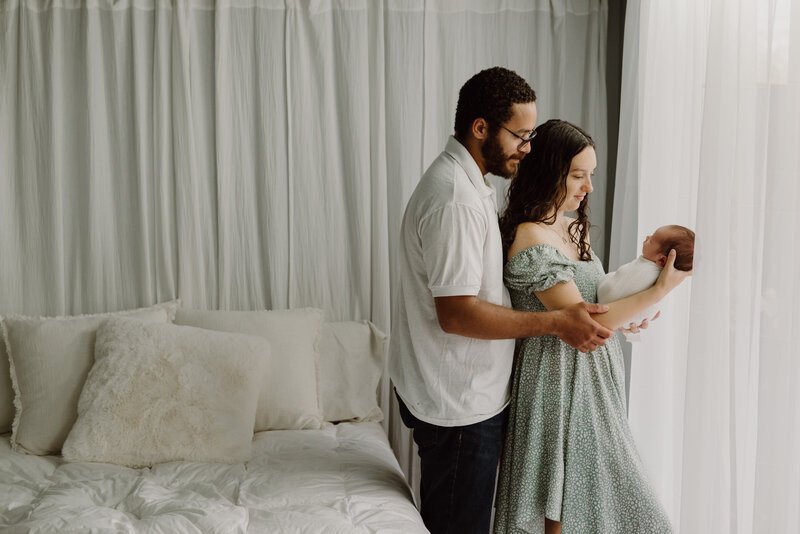Dad and mom standing in front of studio window next to bed looking at baby boy in Annapolis Maryland.