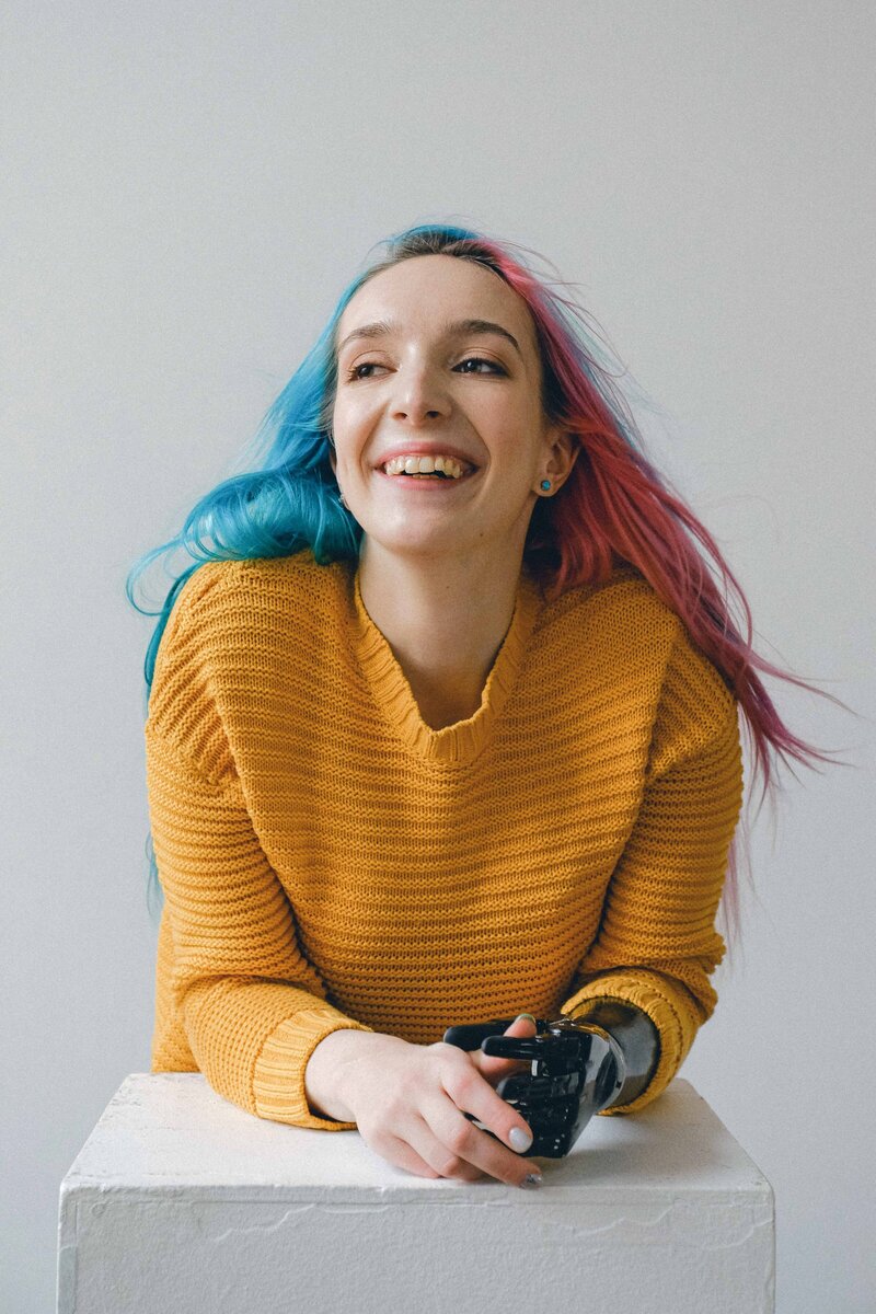 This image shows a young, feminine-presenting white person, leaning forward toward the camera and smiling. They have brightly-colored hair and a black, prosthetic left arm.