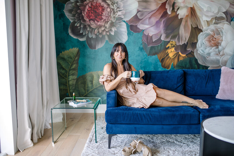 Orlando photographer captures branding photos of woman on a bright blue couch in from=nt of a wall mural of large florals as she holds her coffee mug  for a studio brand session