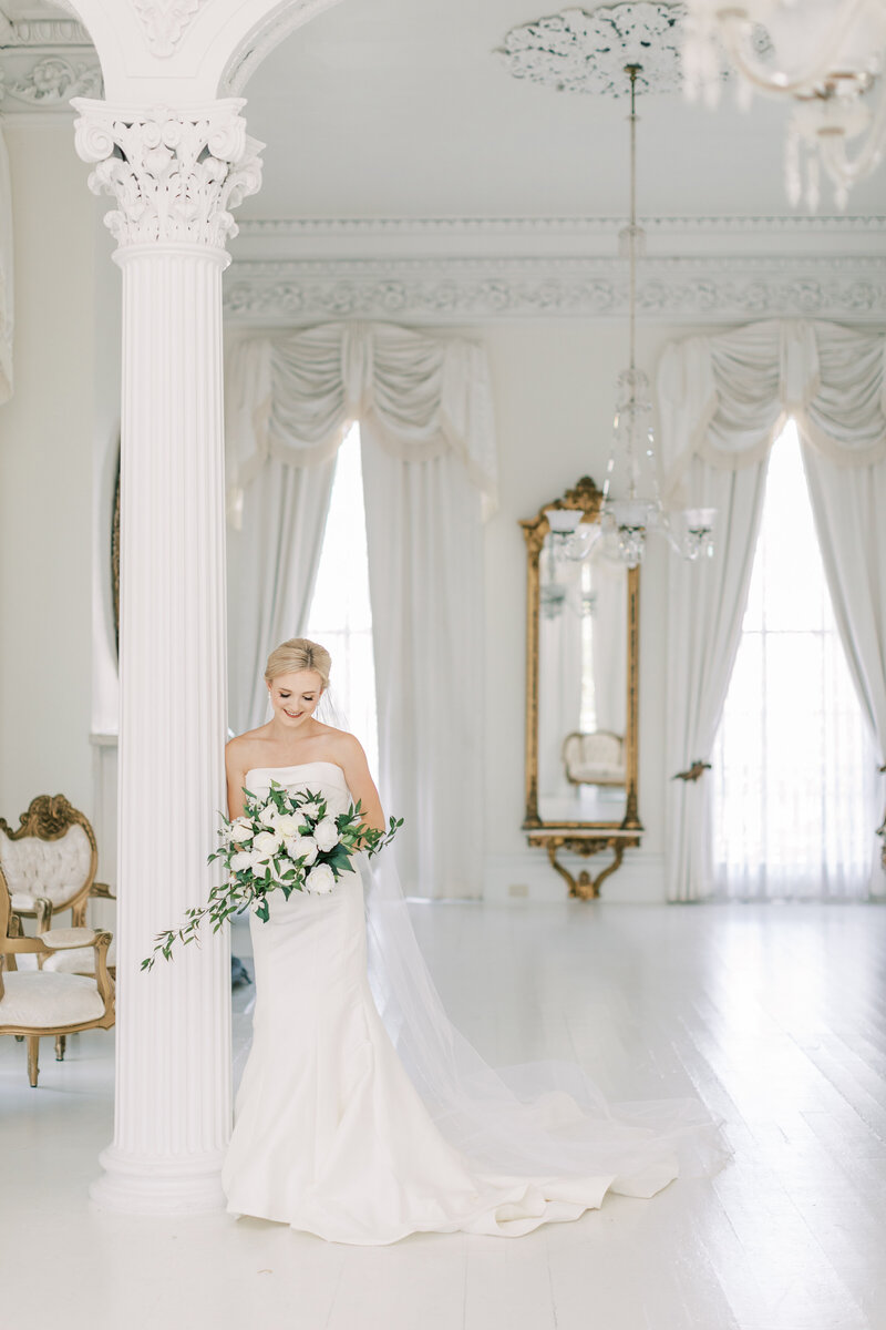 A bride holds her bouquet leaning against a large white column in a white room.