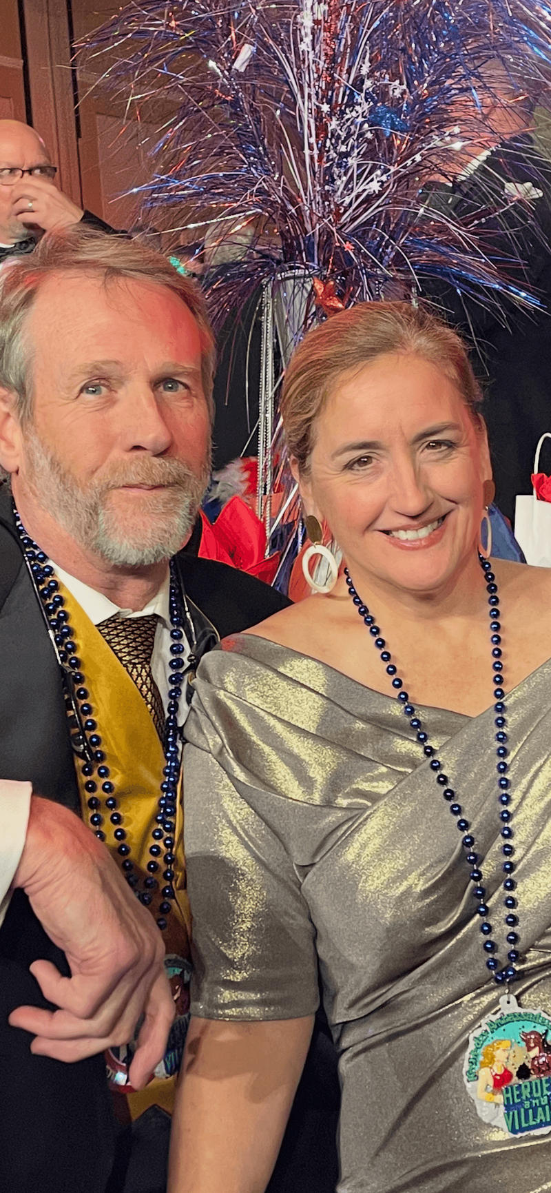 A man in a suit and a woman in a silver dress, both wearing blue bead necklaces, smile at an event with festive decorations and colorful tinsel centerpiece in the background.