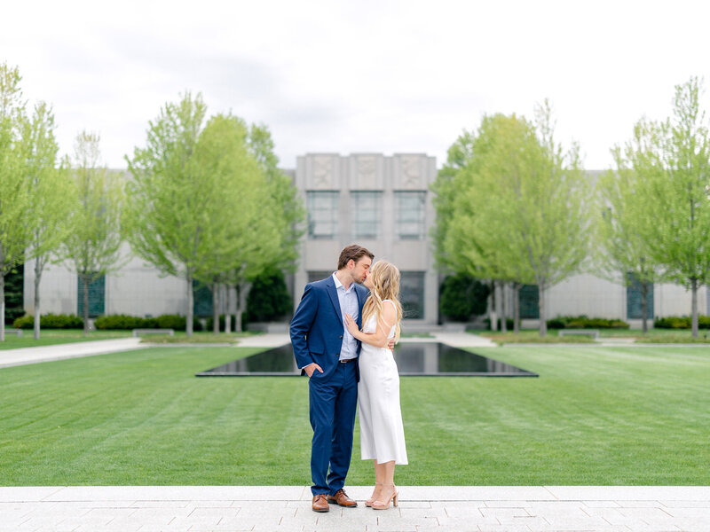 Man in blue suit and girl in white dress kissing in front of a large building and green trees