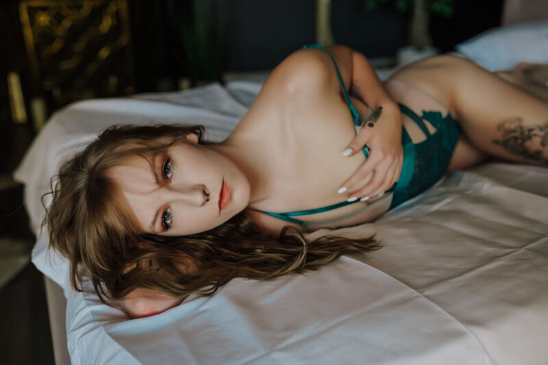 redhead woman in green lingerie with hand on breast laying on white sheets