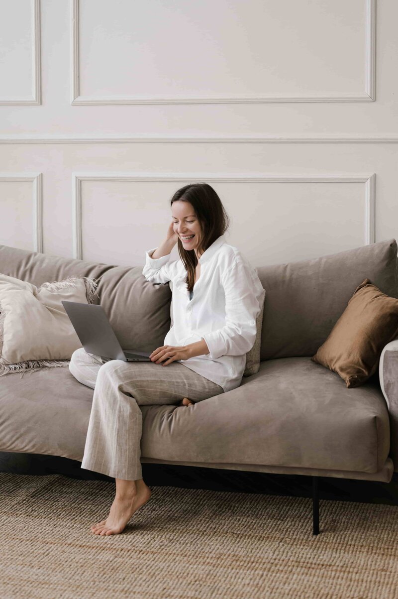 ITN students become a Certified Transformational Nutrition Coach through a completely online program; illustrated by this photo of a woman smiling on a couch while working on her laptop.