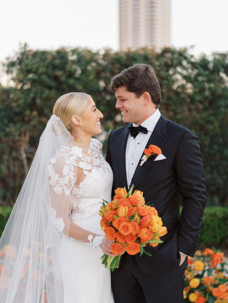 Bride and groom smiling at each other while bride holds orange bouquet