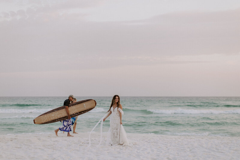 bride stands on beach with surfers behind