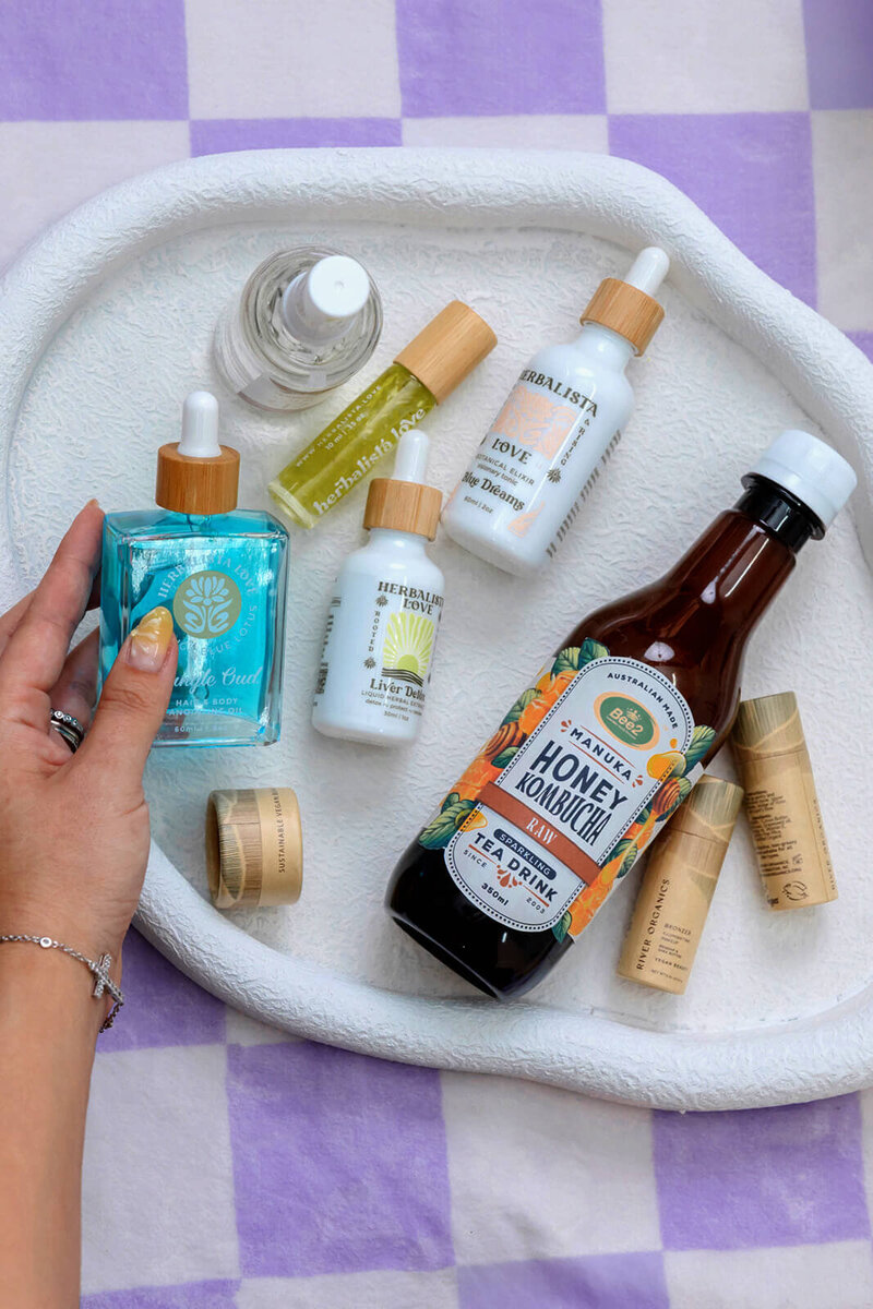 A selection of natural beauty and wellness products displayed on a white tray. The items include Herbalista Love skincare bottles, a blue facial serum, lip balms, and a bottle of Manuka Honey Kombucha. A hand is shown holding the facial serum, highlighting the eco-friendly packaging and natural ingredients of the products, all set against a purple checkered background.