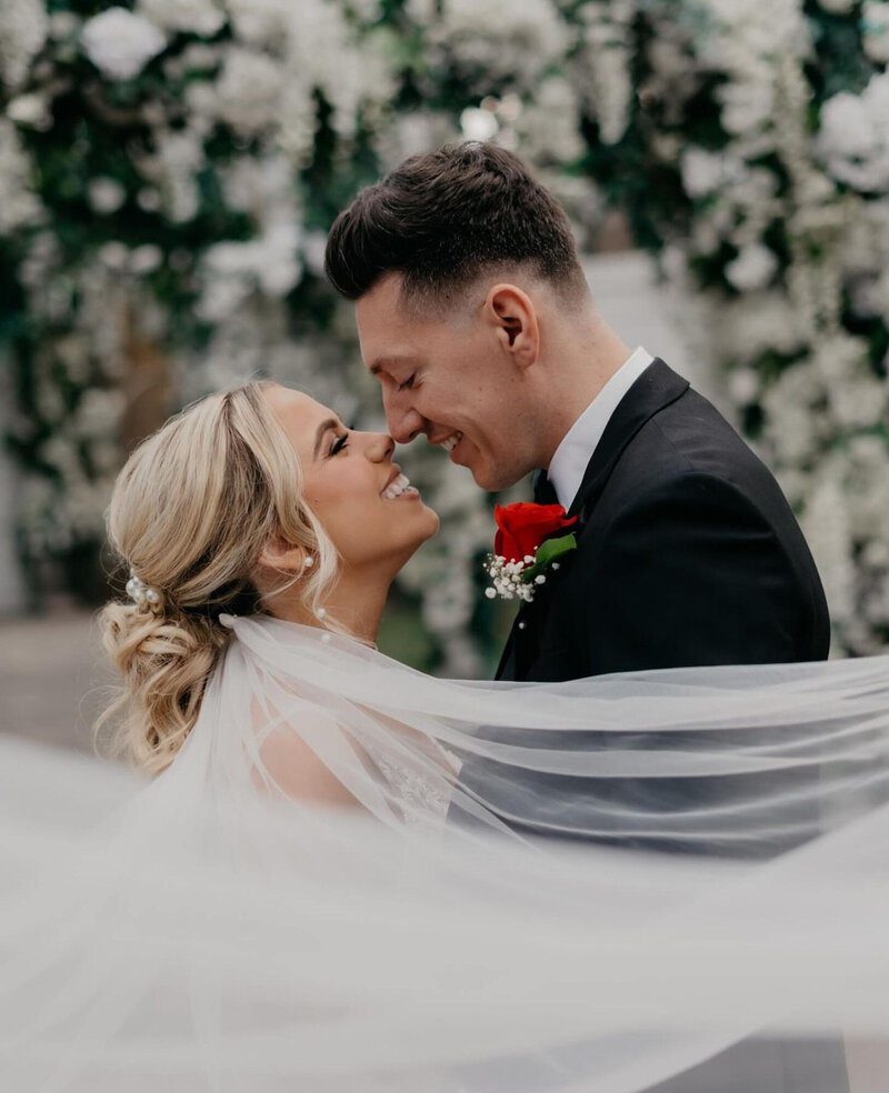 bride veil is gently flowing, and the groom wears a black suit with a red rose boutonniere