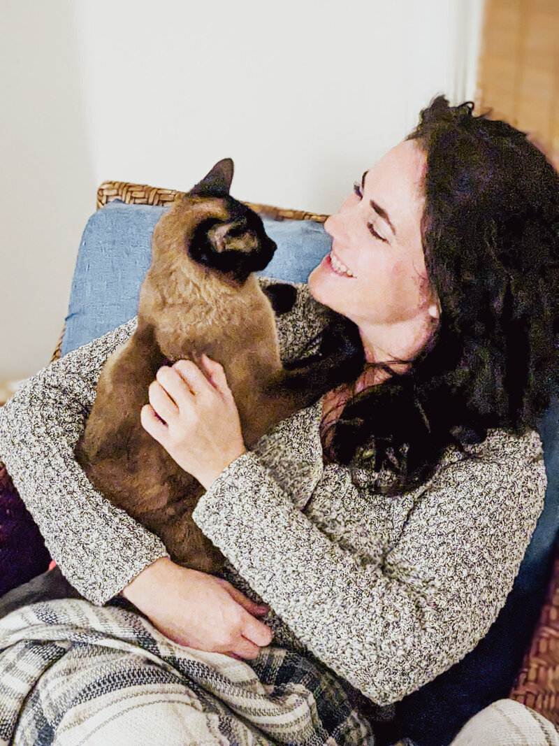 Caroline Busick sitting in a chair holding a Siamese cat in her arms smiling face to face