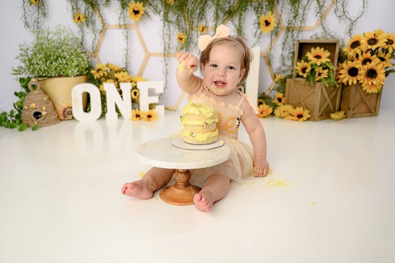 little girl shows off her sharing skills as she sits in her custom "happy bee day" cake smash set up