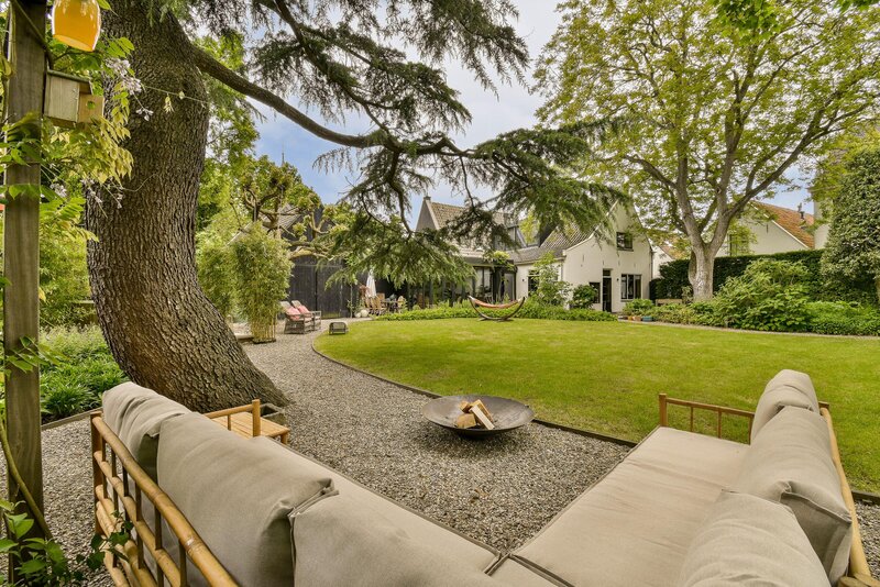 A spacious backyard with a large lawn, mature trees, and comfortable outdoor seating, including a hammock and a fire pit, under a canopy of greenery.