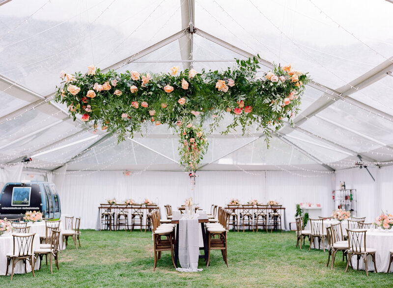 Outdoor tented wedding reception set up. The tent is see-through, and there is a large floral arrangement hanging from the ceiling. The tables and chairs are dark wood with draping white tableclothes.