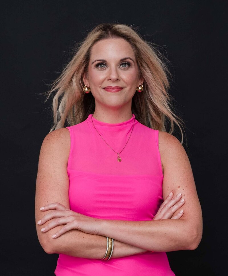 woman with blonde hair and a pink dress with herarms crossed across her chest in a power pose