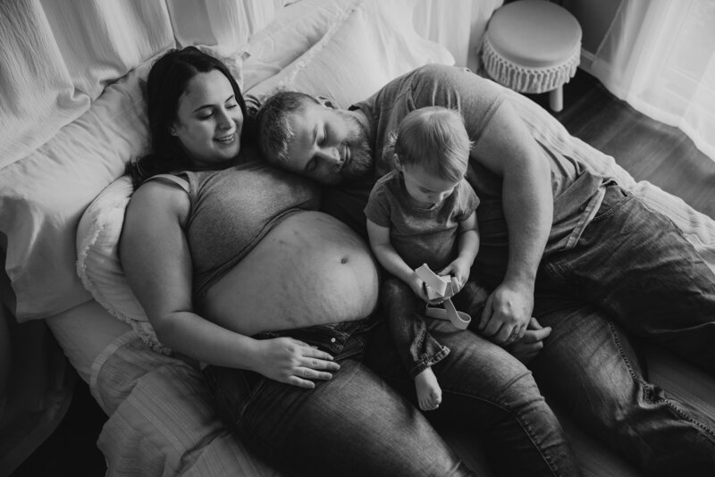 Black and white photo of pregnant mom, dad, and toddler son sitting on bed for Maternity photos in a moody studio in Annapolis Maryland.
