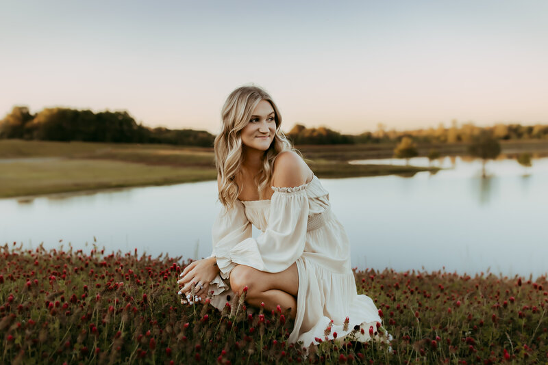 A person in a white off-shoulder dress is kneeling in a field of red flowers near a calm lake during sunset, captured beautifully by Fire Family Photography in Macon GA.