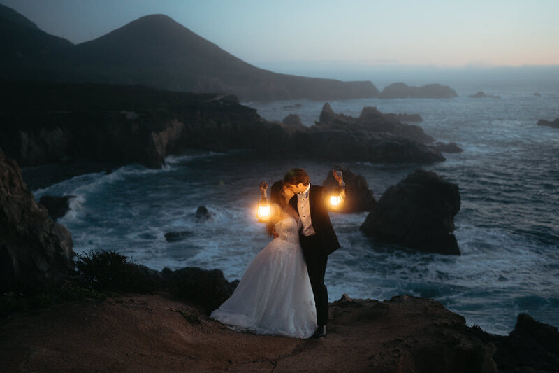 Elopement Photo Ideas | Eloping couple kiss and hold up golden lanterns against a blue ocean with Big Sur coastline behind them