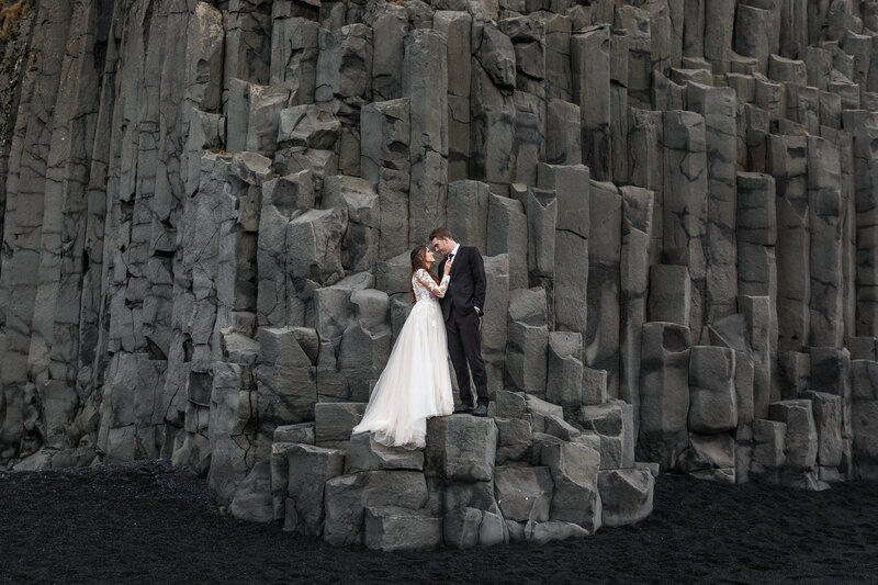 A bride and groom look at each other on basalt columns on a black sand beach in Iceland.