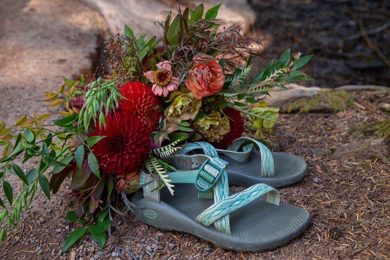 Bridal bouquet with red and green florals resting on teal Chaco sandals on a forest trail.