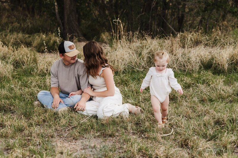 Lubbock family sitting on the grass, couple is cuddled up and their little girl is walking around them exploring. Momma and daughter are dress in cream outfits and dad is in denim jeans and gray t shirt.